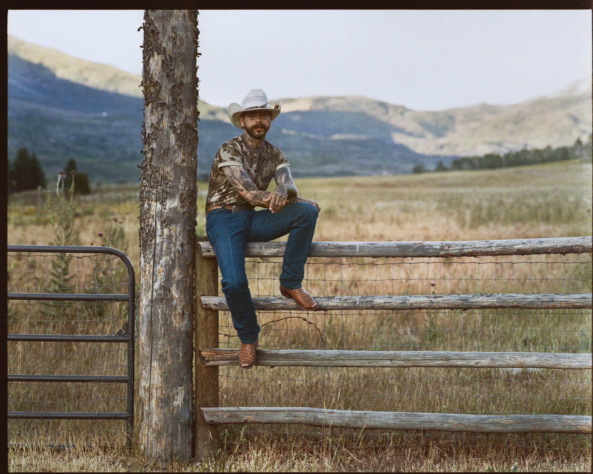 Post Malone seated on a wooden fence wearing a camo t-shirt and blue jeans in an outdoor ranch setting.