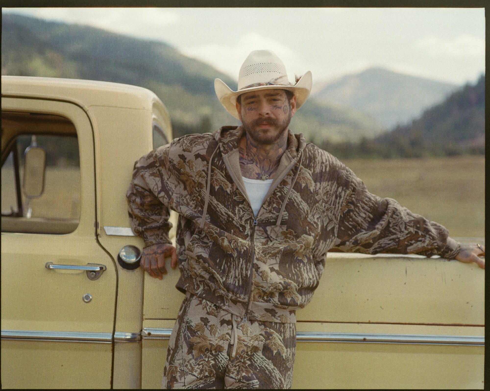 Post Malone leaning against a vintage yellow pickup truck wearing camo print hoodie and cowboy hat outdoors.