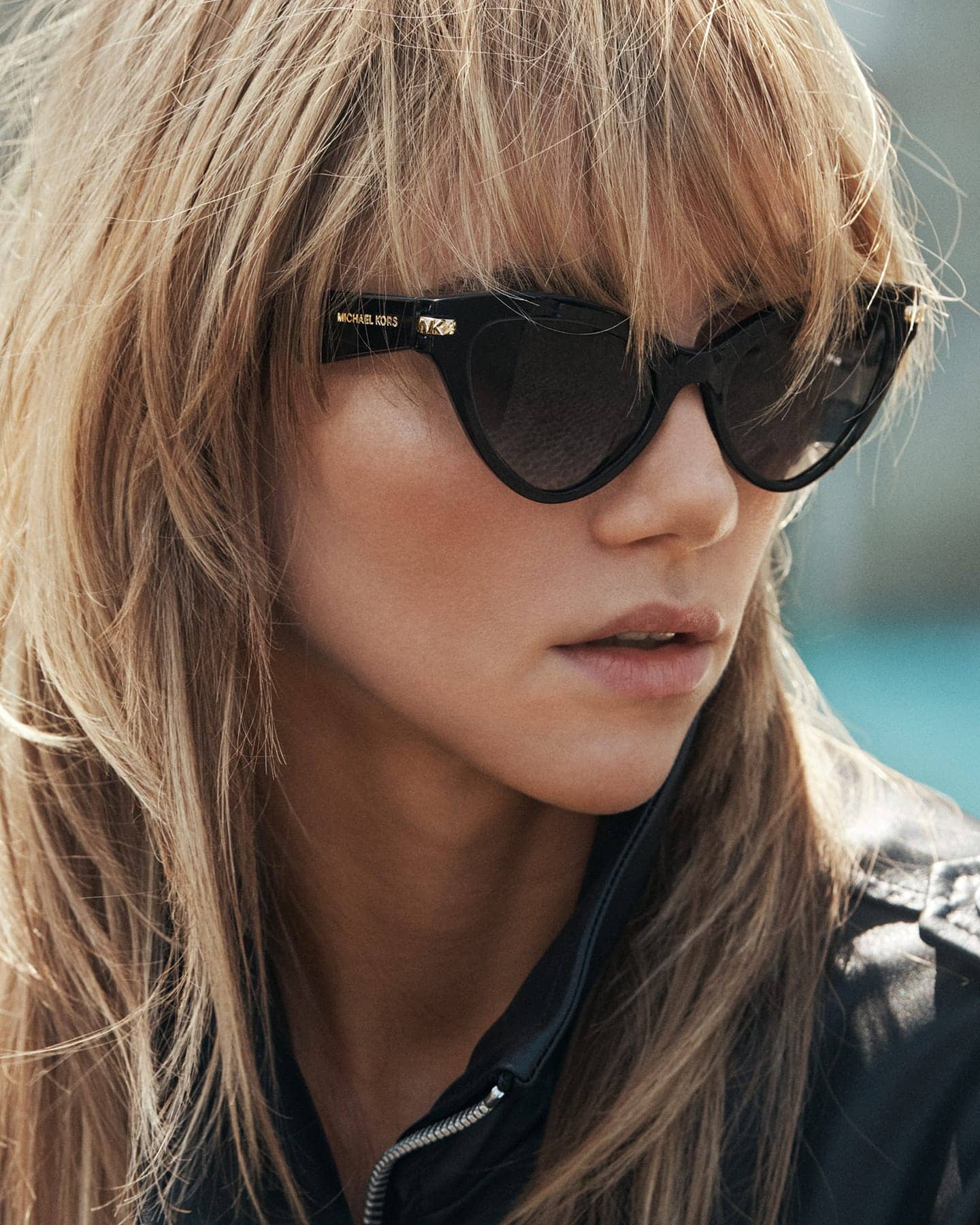 Close-up portrait of a blonde woman wearing black cat-eye sunglasses and a dark zipped jacket for Michael Kors Fall 2025 ad campaign.