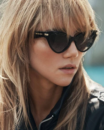 Close-up portrait of a blonde woman wearing black cat-eye sunglasses and a dark zipped jacket for Michael Kors Fall 2025 ad campaign.