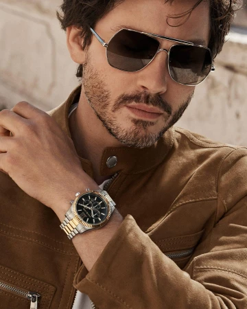 Man wearing sunglasses and a brown jacket with a wristwatch on his wrist