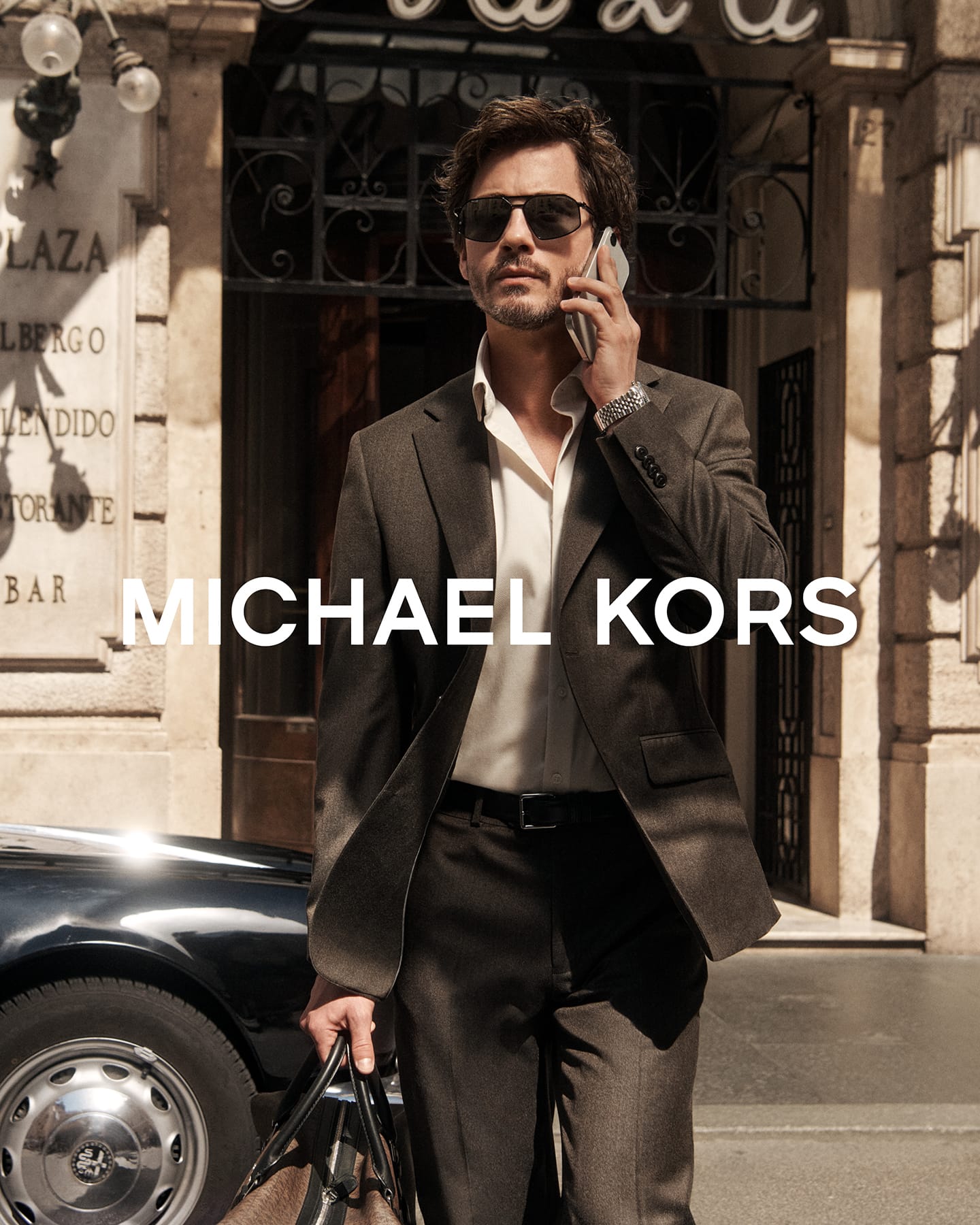 Logan Lerman in a dark suit jacket holding a phone outside Grand Hotel Plaza in Rome for Michael Kors