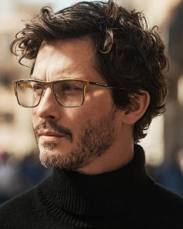 Close-up portrait of a man wearing gold-framed eyeglasses and a black turtleneck sweater outdoors