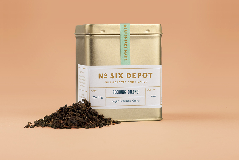 Gold tea tin with No. Six Depot label and loose dark oolong tea leaves on a warm peach background