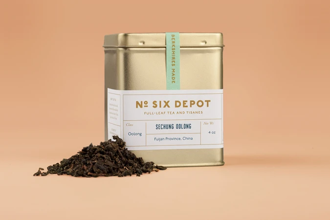 Gold tea tin with No. Six Depot label and loose dark oolong tea leaves on a warm peach background