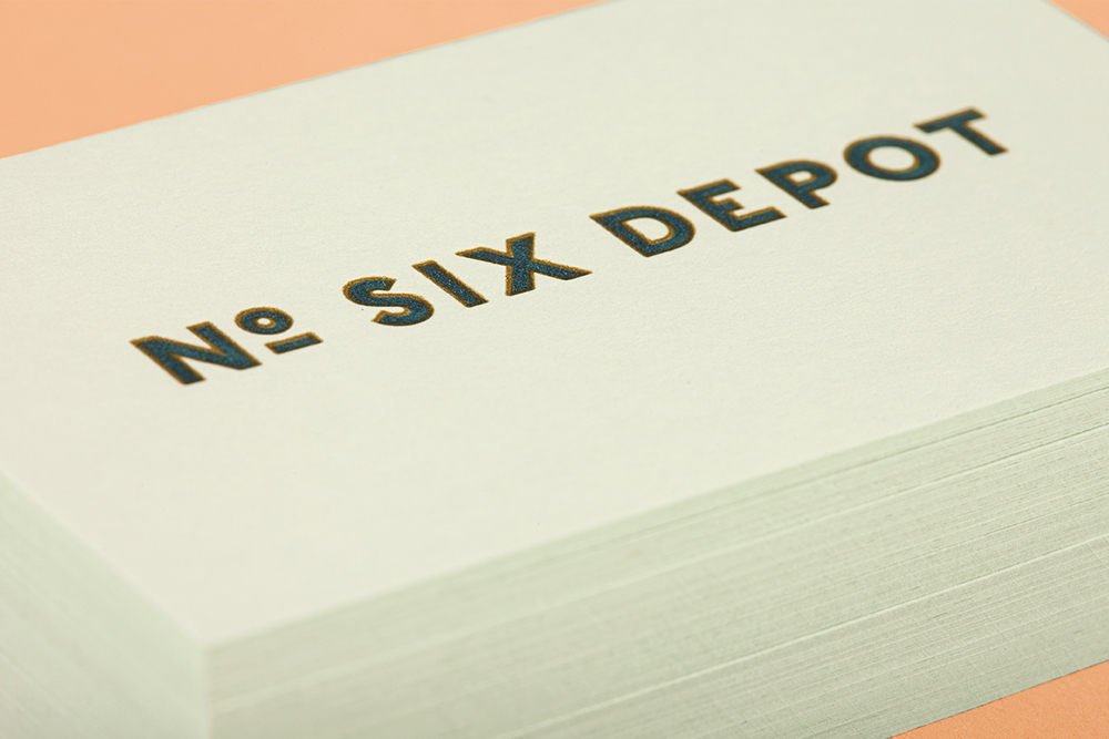 A stack of white business cards featuring embossed dark text reading NO SIX DEPOT on a peach surface