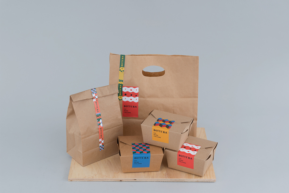 Brown paper bags and take-out boxes with colorful branded labels arranged on a wooden board against a light grey background.