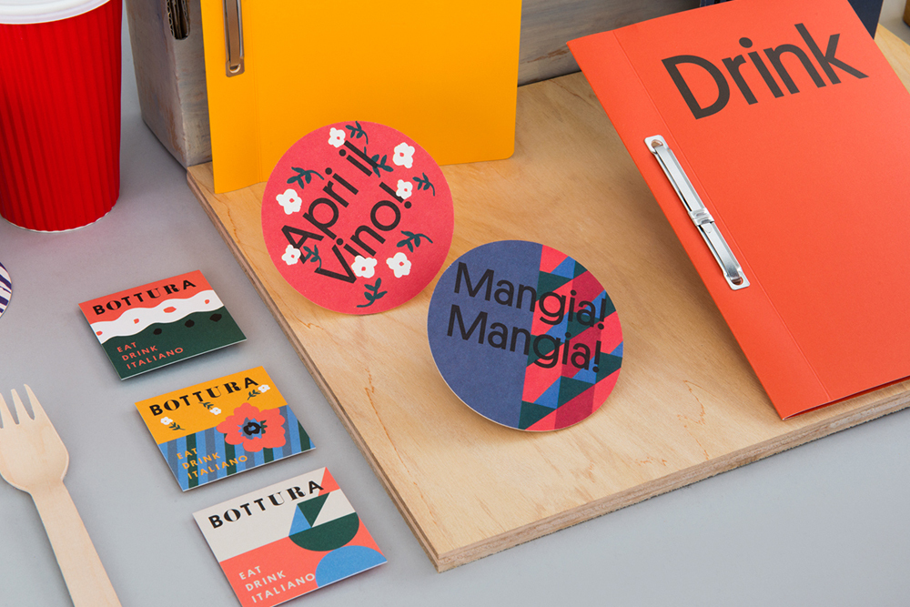 Bottura brand identity stationery flat lay with red folder, colourful coasters and wooden fork on a grey surface.