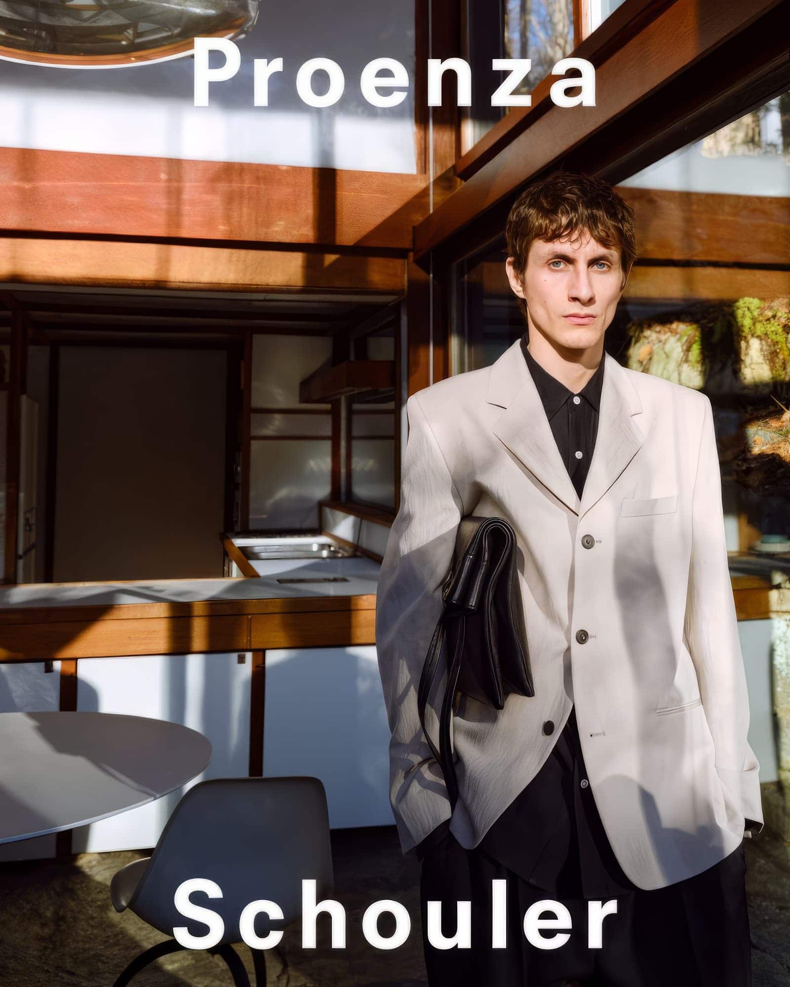 Man wearing a light grey blazer holding a black handbag inside a modern architectural interior