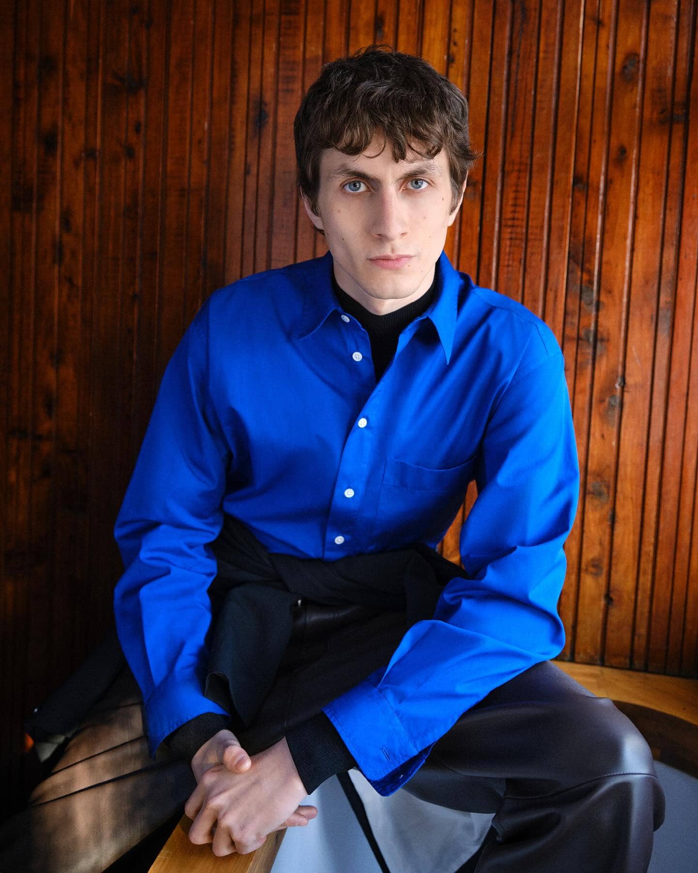 Man wearing a vibrant blue button-up shirt and dark trousers seated against a vertical wooden panel background.