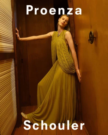 Model in pleated olive dress leaning against a wooden door frame in the Proenza Schouler Fall 2025 campaign.
