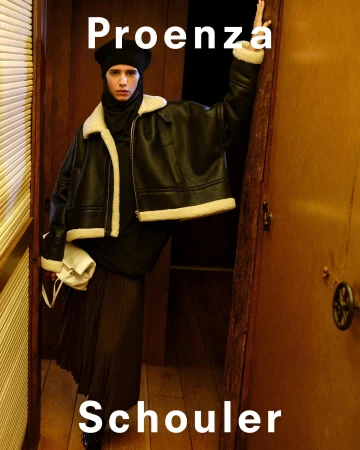 Model in dark beret and black shearling-lined jacket leaning in a wooden interior doorway holding a white handbag.