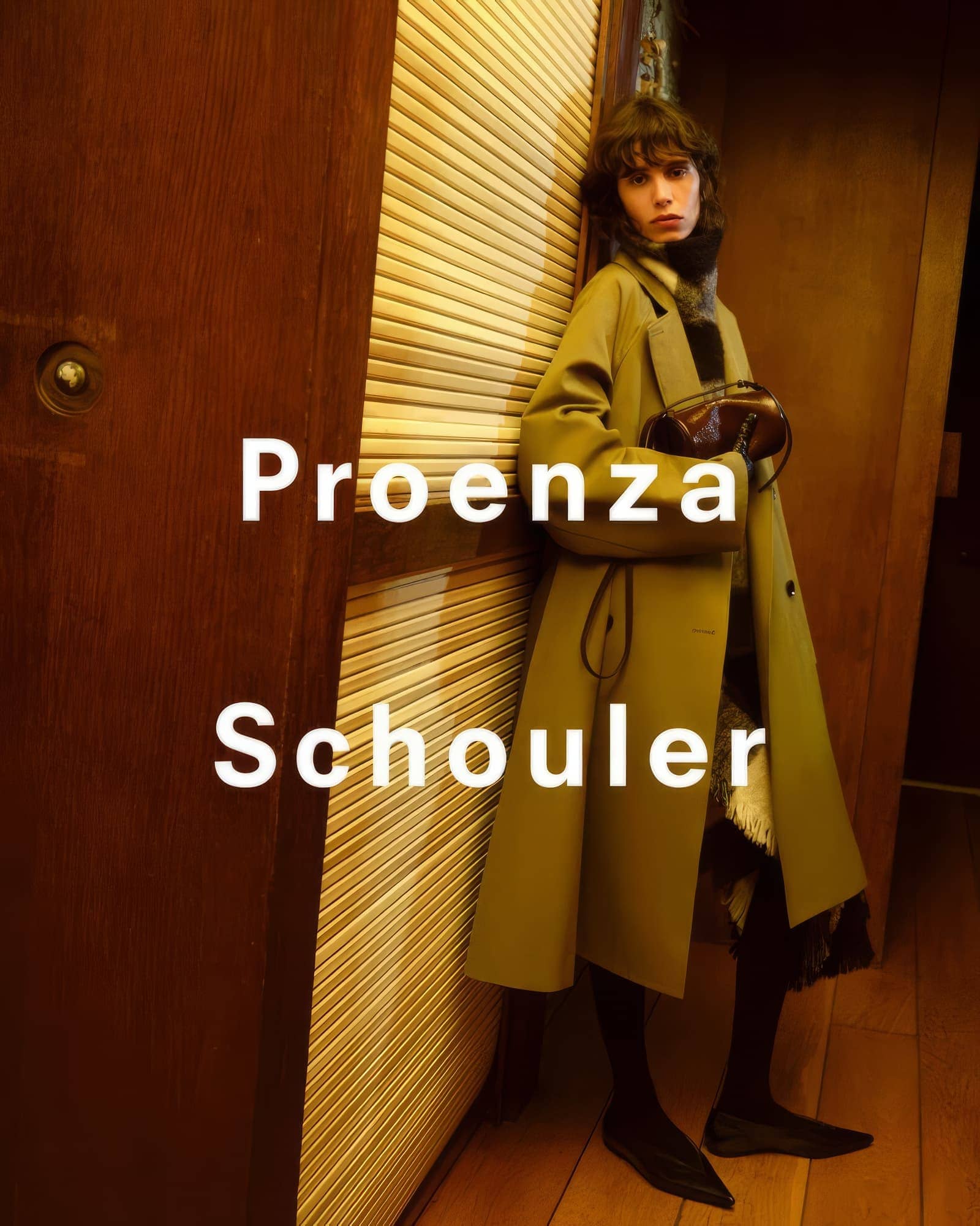 Model in olive green coat leaning against wooden slats with dark interior background