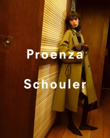 Model in olive green coat leaning against wooden slats with dark interior background