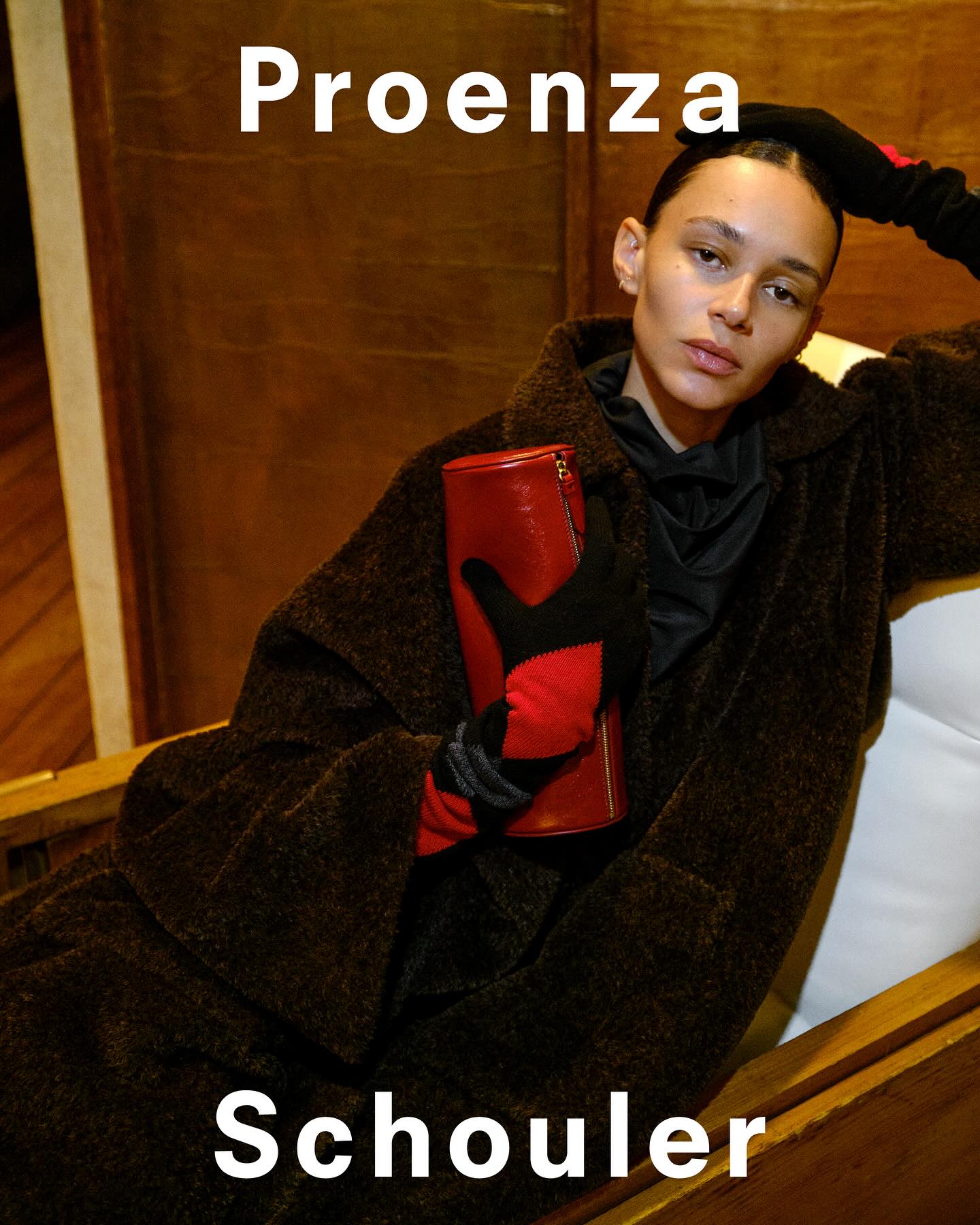 Model wearing dark textured coat and red gloves holding a cylindrical leather clutch for Proenza Schouler