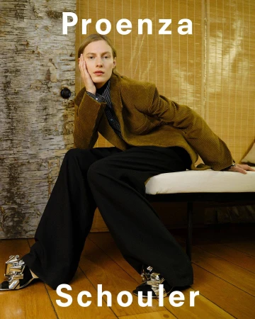 Model seated wearing an olive blazer and black trousers against a textured wooden background for Proenza Schouler