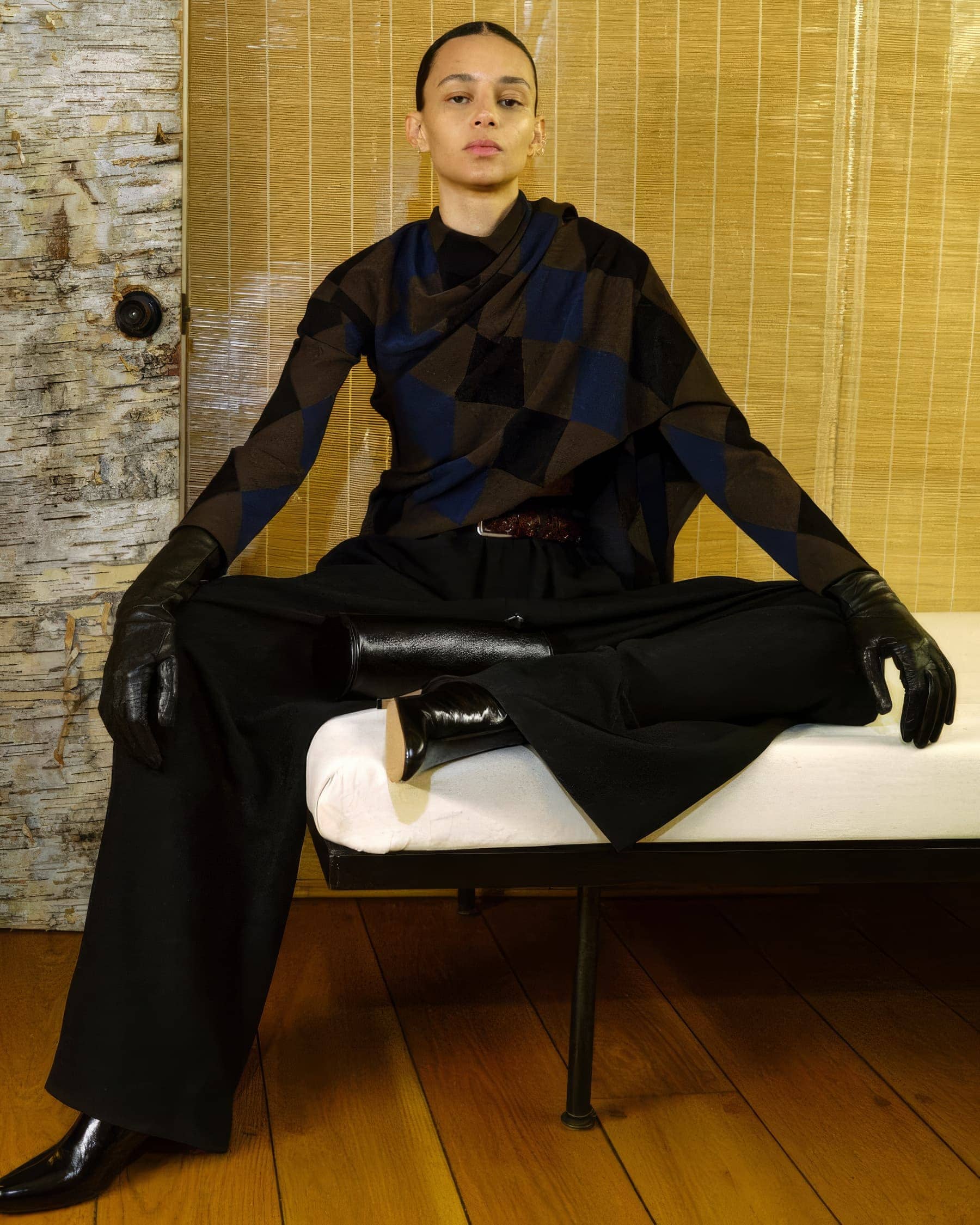 Model seated cross-legged wearing a patterned argyle top, wide black trousers, and black leather gloves
