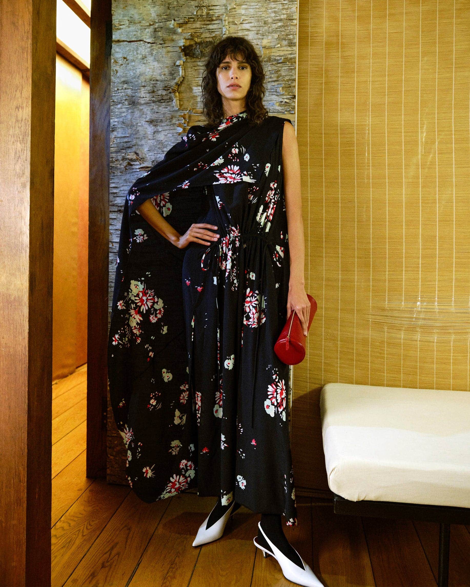 Model in a black floral dress with matching draped cape holding a red clutch against a textured wall