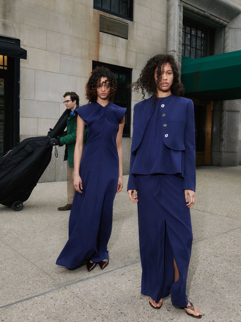 Two fashion models in blue dresses pose on a city street with a man pulling luggage in the background, showcasing modern fashion.