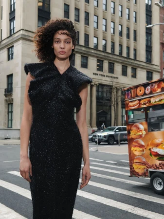 Woman in a black dress stands on a crosswalk in New York City, with a food cart and a building marked “New York University” in the background.