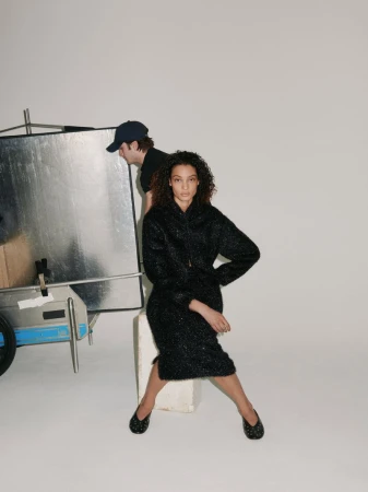 A woman in a dark, textured skirt suit sits posing in a studio, with a lighting assistant holding a reflector in the background.