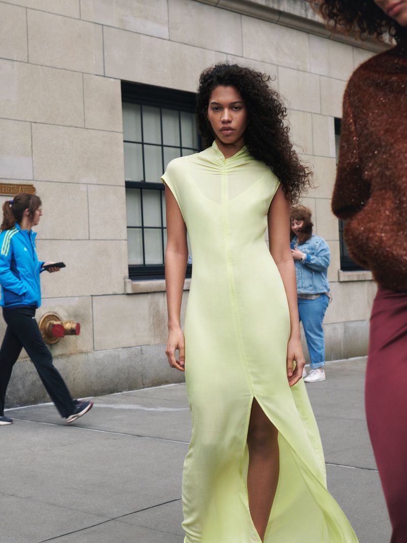 A woman in a long, light green dress stands on a city street with pedestrians in the background and a neutral-colored building behind her.