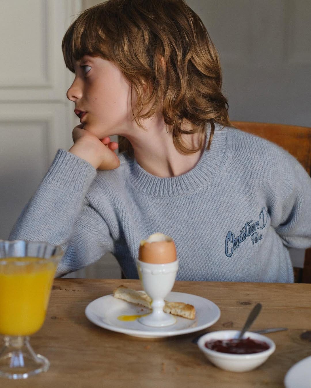 Young child in a grey sweater seated at a wooden table with a boiled egg and juice for Dior Kids Spring 2025 campaign.