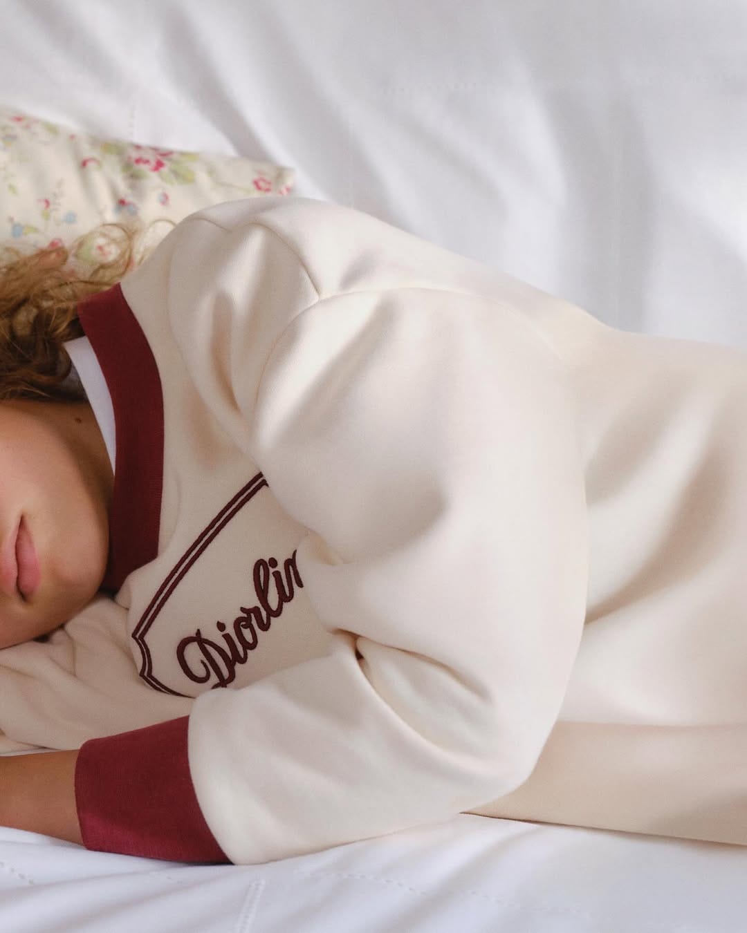 Child resting on white bedding wearing a cream sweatshirt with burgundy trim for Dior Kids