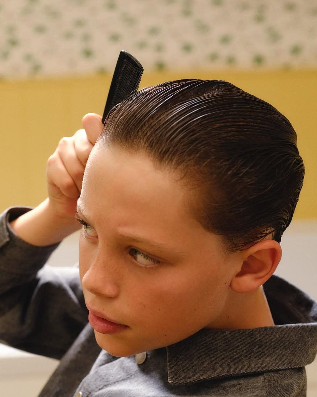 Young child combing dark slicked-back hair while wearing a grey collared shirt, captured in a close-up portrait.
