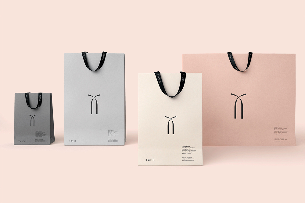 Four paper shopping bags in varying sizes and pastel tones with black handles and minimal branding arranged on a pale pink backdrop