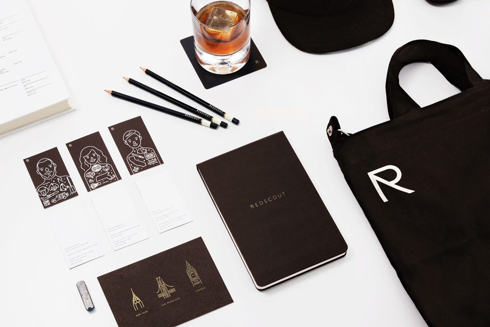 Overhead flat lay of Redscout brand identity collateral including a notebook, tote bag, pencils, cards, and a glass of whiskey.