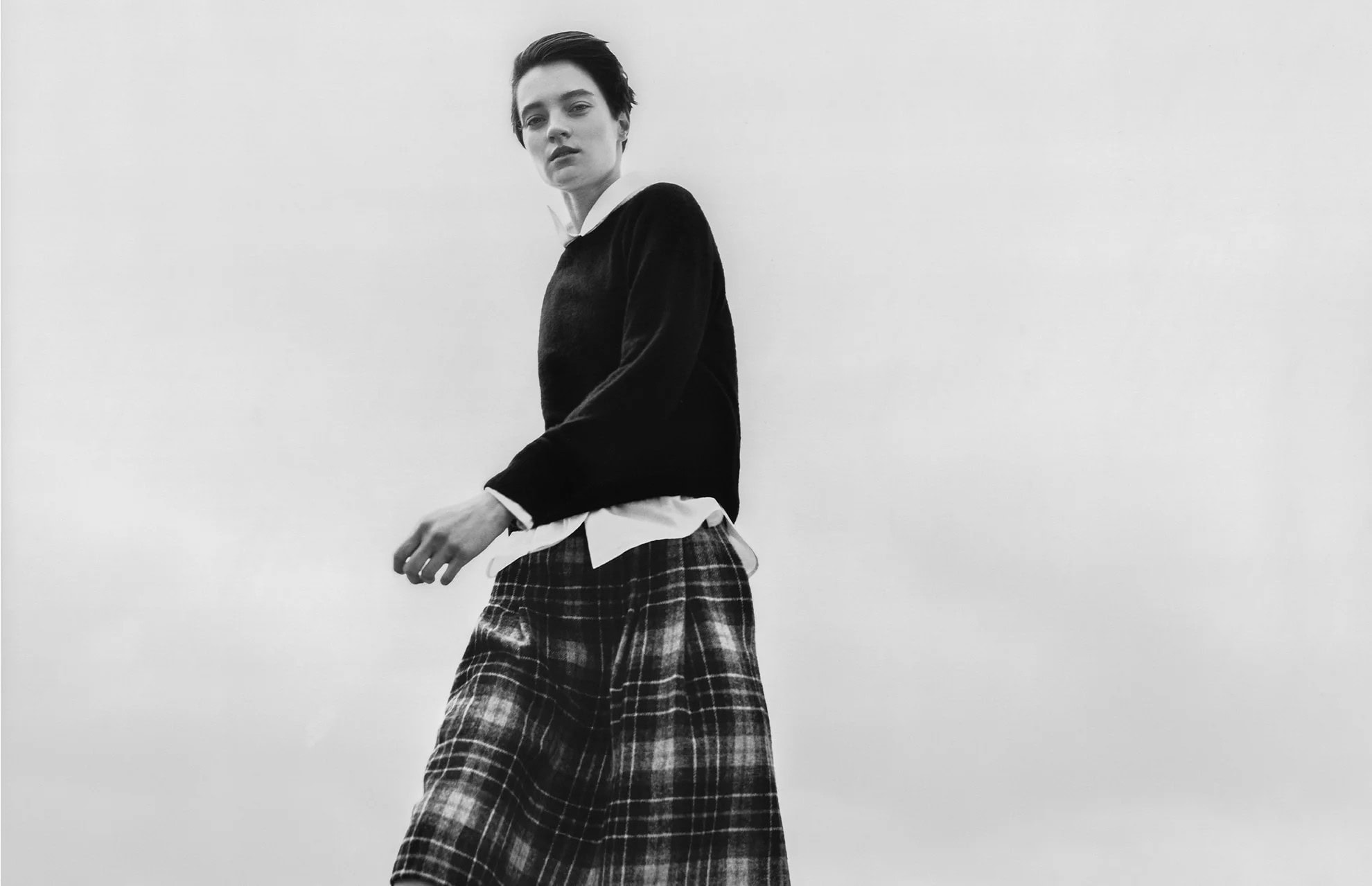 Model in dark sweater and plaid skirt against a pale overcast sky for Margaret Howell campaign.