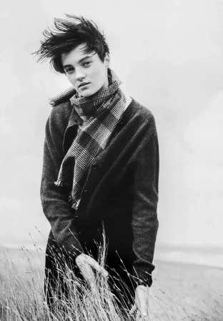 Model with wind-swept dark hair wearing a patterned scarf and knit sweater outdoors against a coastal background