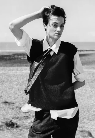 Model with dark short hair wearing a sweater vest, white shirt, and tie against an outdoor coastal background.