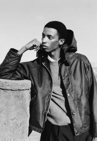 Black man wearing a dark waxed jacket and knitted sweater leaning against a stone wall outdoors