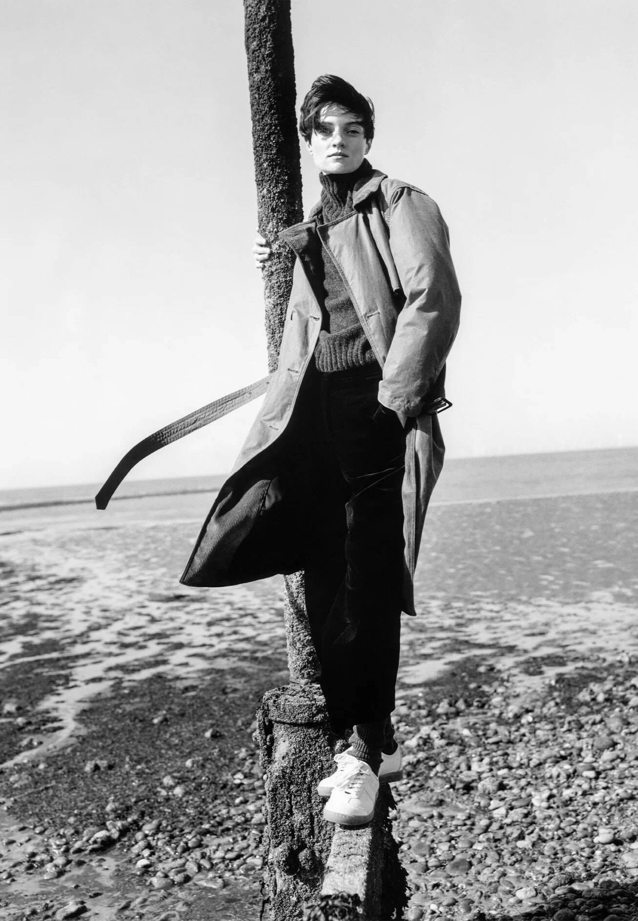 Model in trench coat and dark trousers leaning against a wooden post on a pebble beach