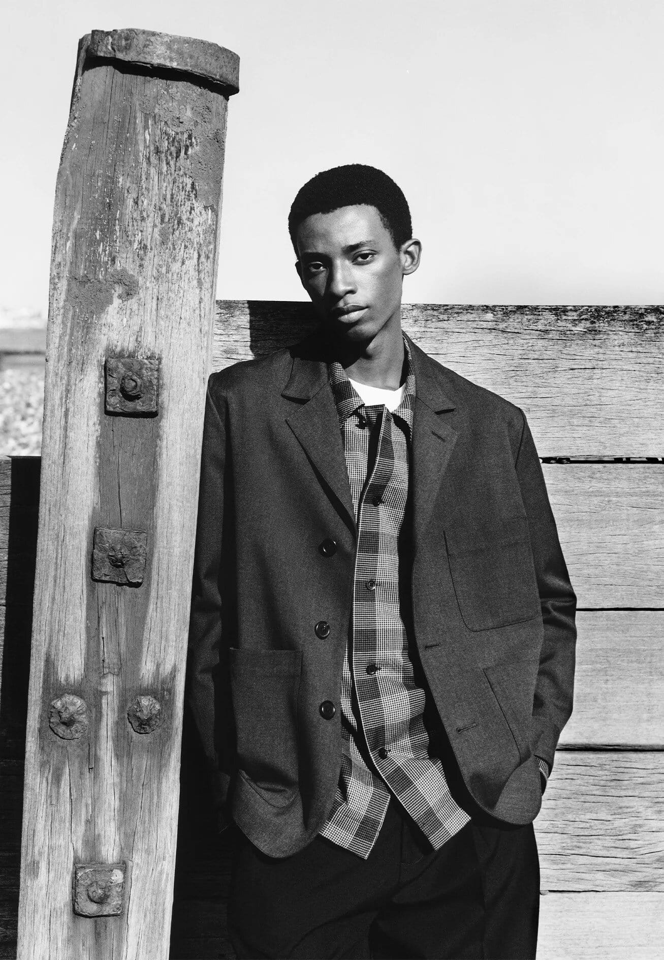 Black man leaning against a weathered wooden pier post wearing a dark blazer over a plaid shirt