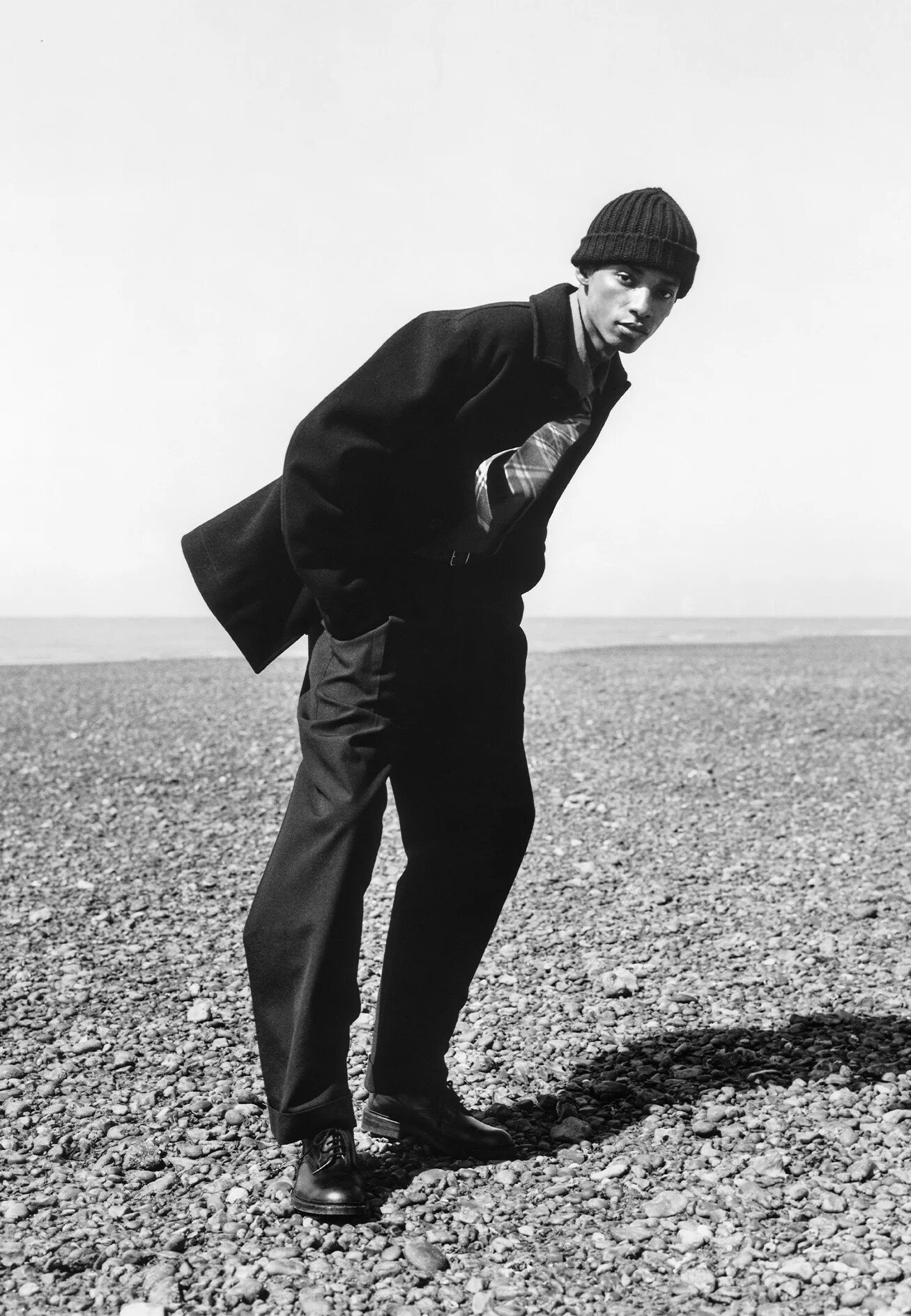 Black and white portrait of a man wearing a beanie and dark jacket standing on a pebble-covered coast by the water.