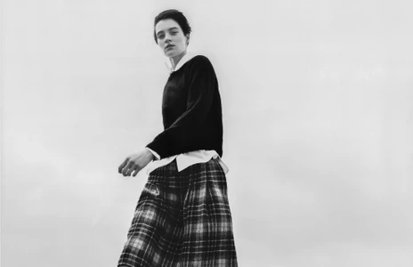 Model in dark sweater and plaid skirt against a pale overcast sky for Margaret Howell campaign.