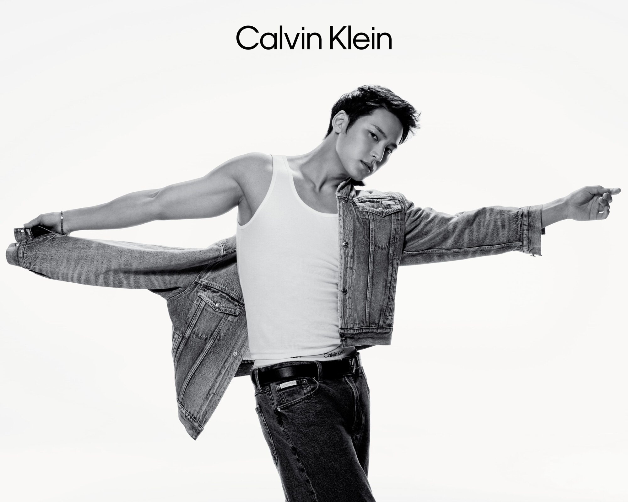 Black and white portrait of Mingyu wearing a white tank top and denim jeans, taking off a denim jacket against a white background.