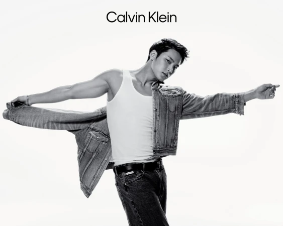 Black and white portrait of Mingyu wearing a white tank top and denim jeans, taking off a denim jacket against a white background.