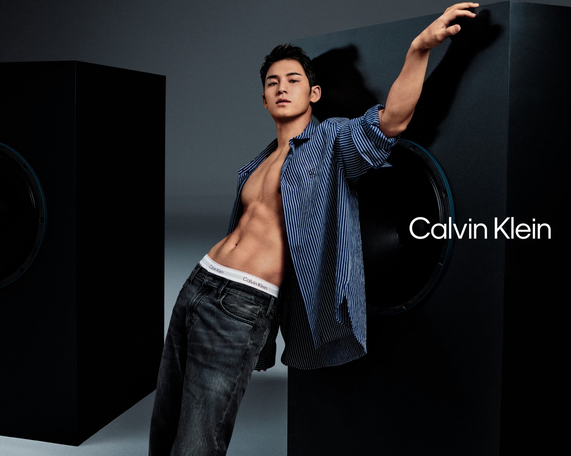 Mingyu posing for Calvin Klein in a blue striped unbuttoned shirt and dark jeans against a dark studio background