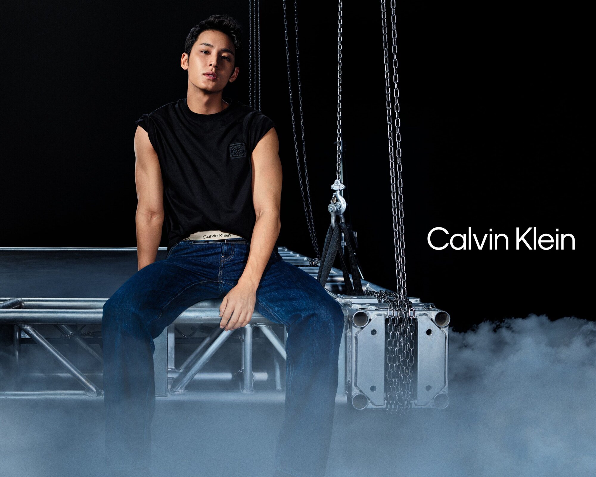 Mingyu wearing black t-shirt and blue denim jeans seated on metallic scaffolding against dark background with smoke