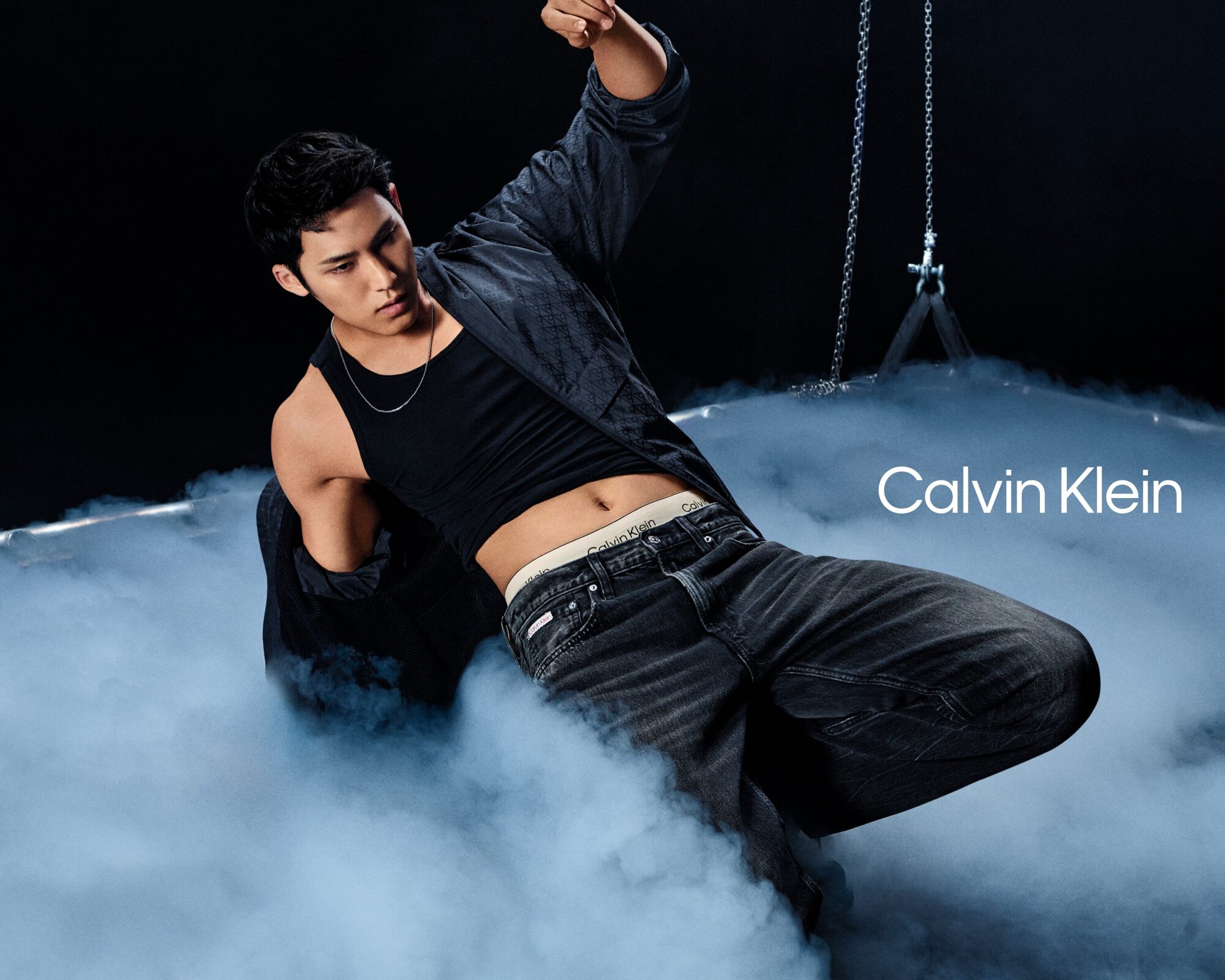Man wearing a black tank top and dark jeans posing amidst low-lying fog with Calvin Klein branding visible.