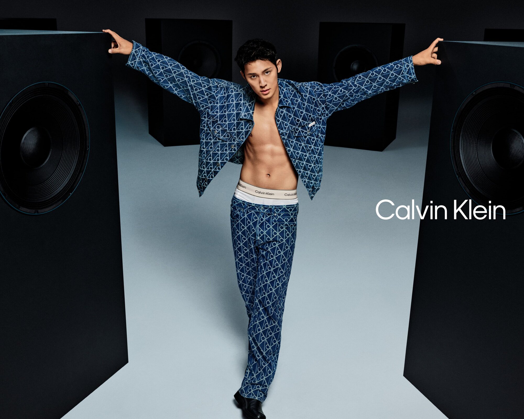 Man wearing monogram denim jacket and jeans standing arms outstretched between large speakers for Calvin Klein ad