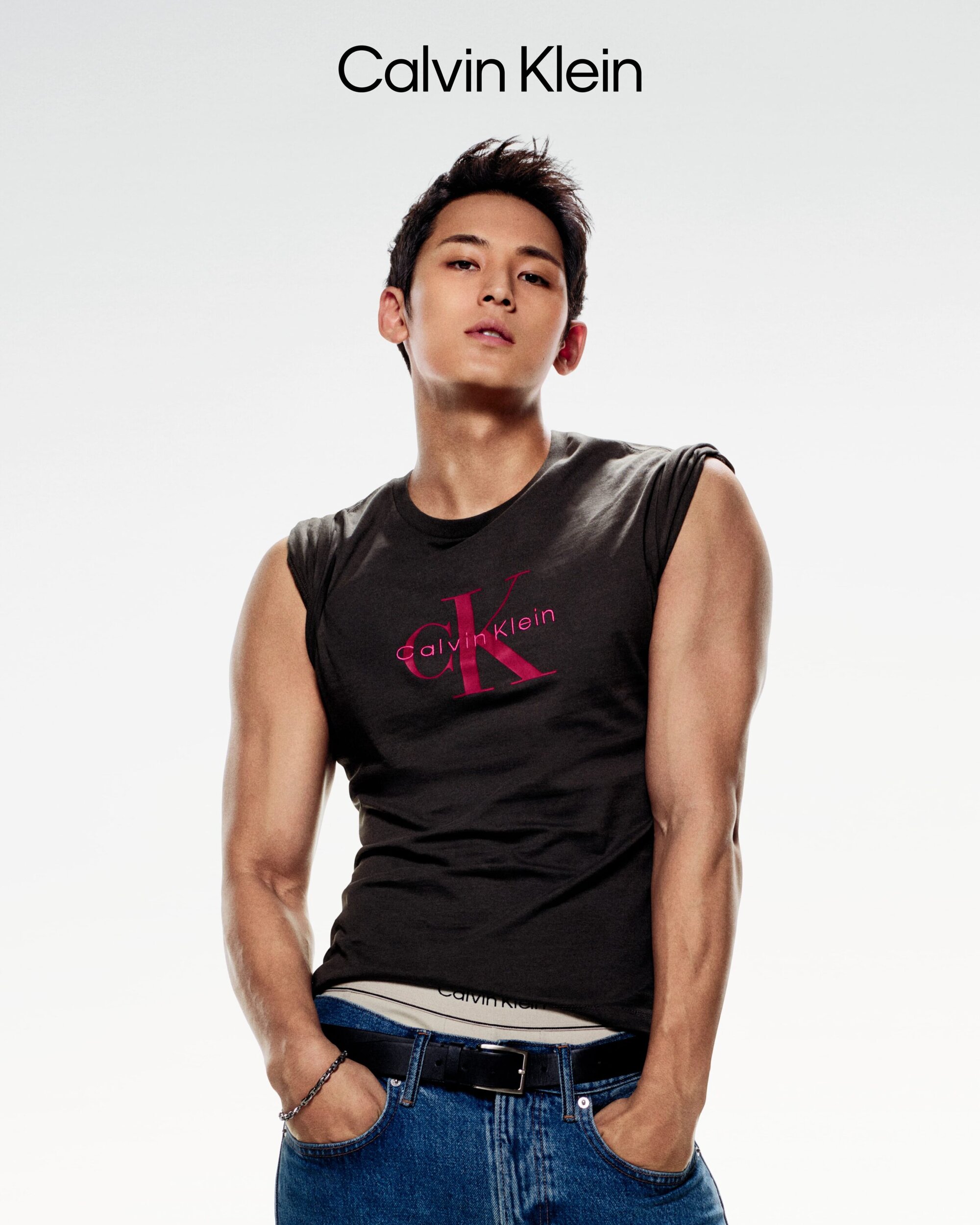 Man wearing a black sleeveless t-shirt and blue jeans posing against a white background for Calvin Klein.