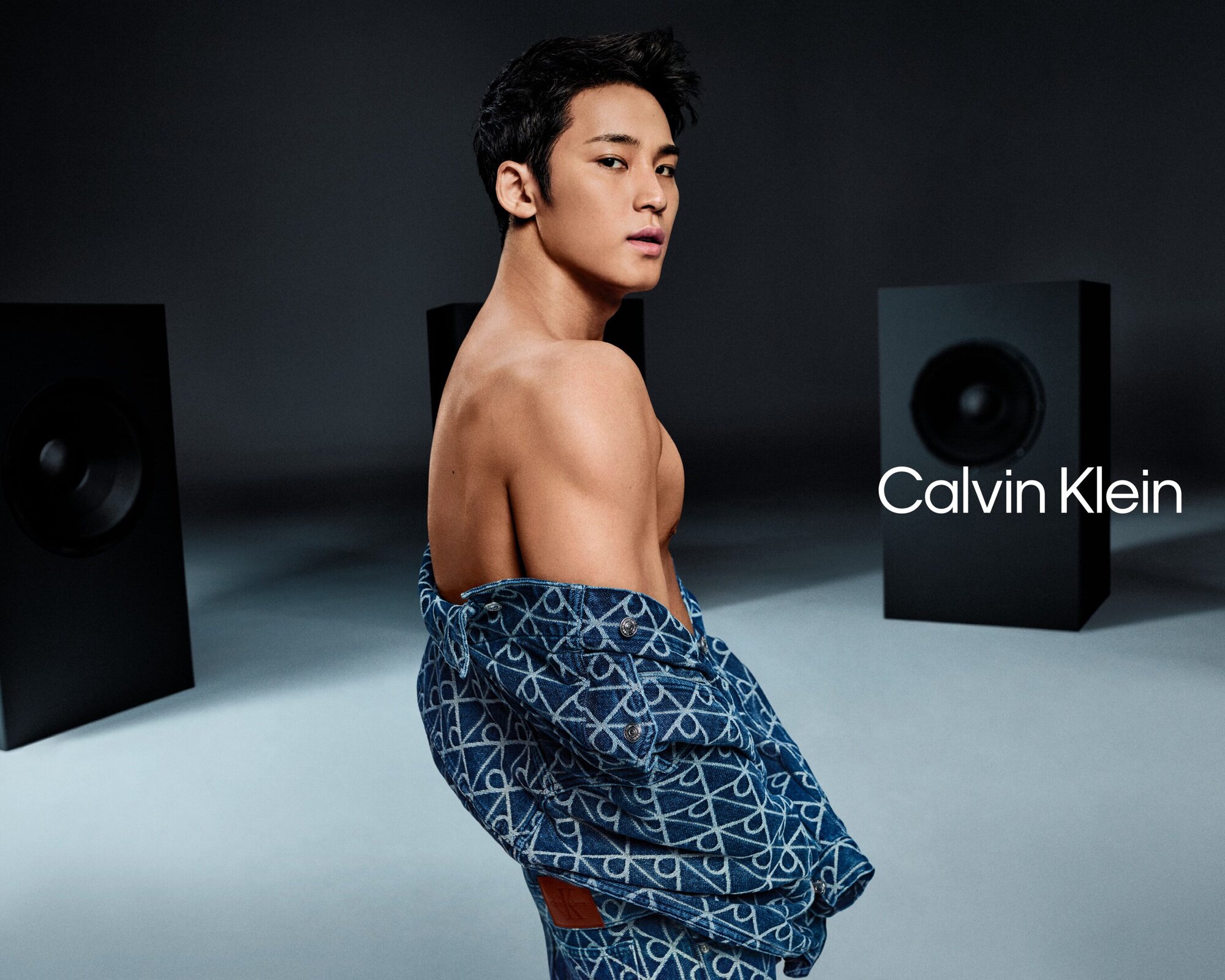 Mingyu posing shirtless wearing patterned denim jacket against a grey studio background with large speakers for Calvin Klein
