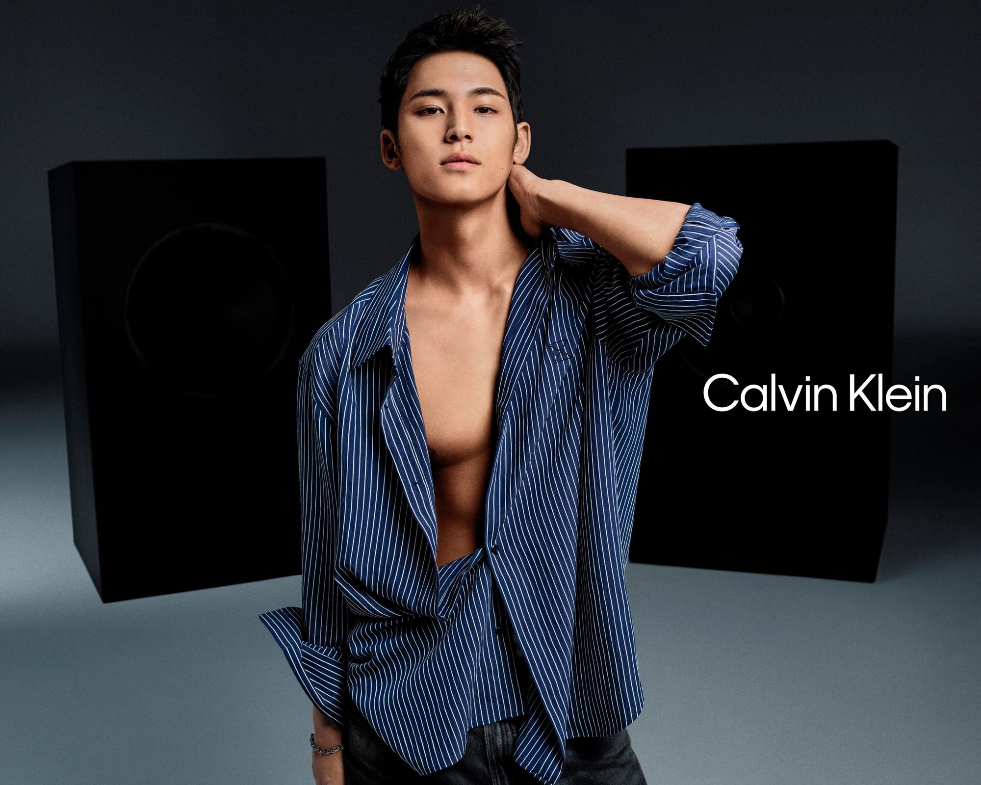Man wearing a blue striped unbuttoned shirt against a grey studio background with large black speakers for Calvin Klein