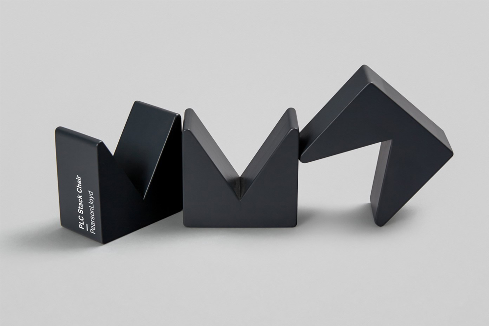 Three dark geometric sculptural objects arranged on a smooth light grey background with visible white typography on the left piece.
