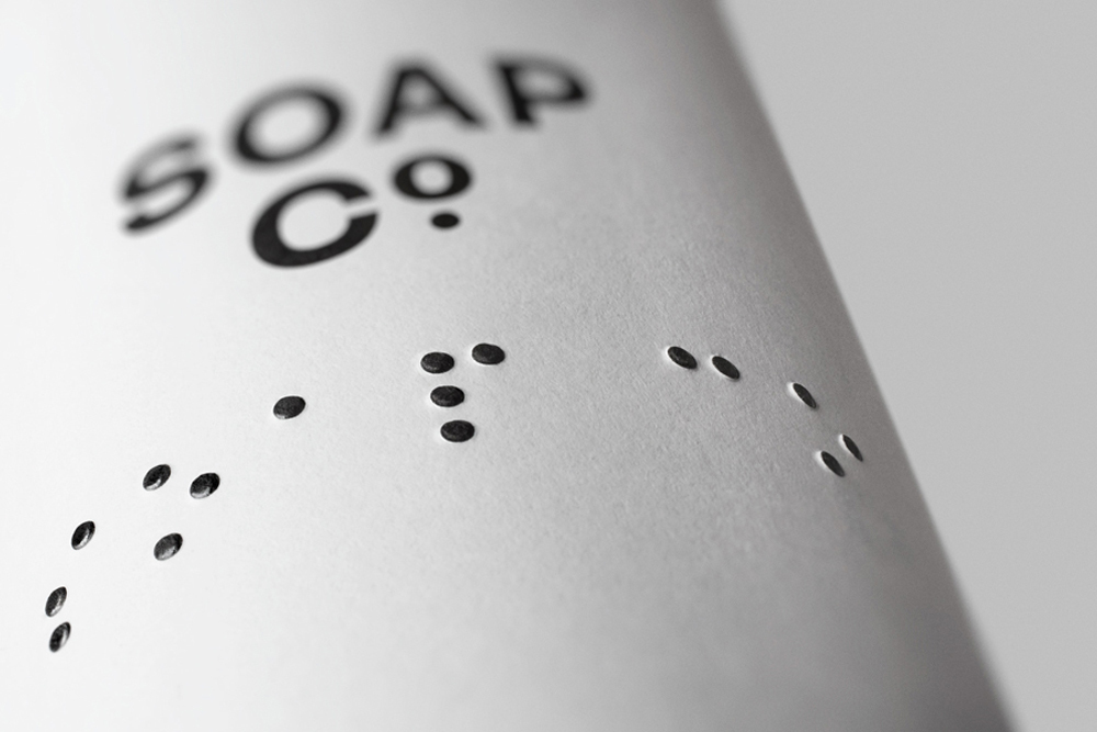 Close-up of The Soap Co. packaging showing black typography and debossed tactile braille dots on textured paper.